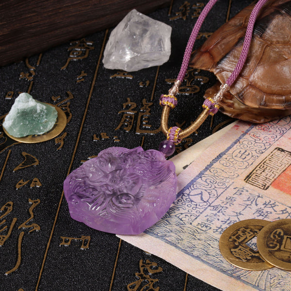 Side perspective view of the deep purple quartz fox pendant laid flat across a traditional hand-painted Chinese character scroll next to a pure raw clear quartz chunk on a dark table surface.