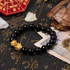 Perspective view of the polished black volcanic glass bead bracelet with a golden Pixiu amulet, arranged on a vintage manuscript near a traditional turtle shell and ancient cash coins.