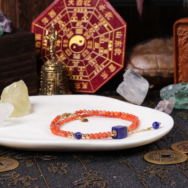 A delicate dual-loop gemstone bracelet featuring vibrant orange-red Southern Red Agate micro-beads and a focal cube-shaped royal blue lapis lazuli stone, displayed on a white ceramic plate within a Feng Shui altar setup.