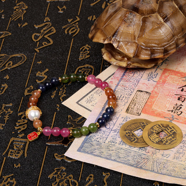 Side perspective view of the colorful rainbow tourmaline talisman bracelet laid over a traditional Chinese manuscript scroll next to ancient copper coins and a textured turtle shell artifact.