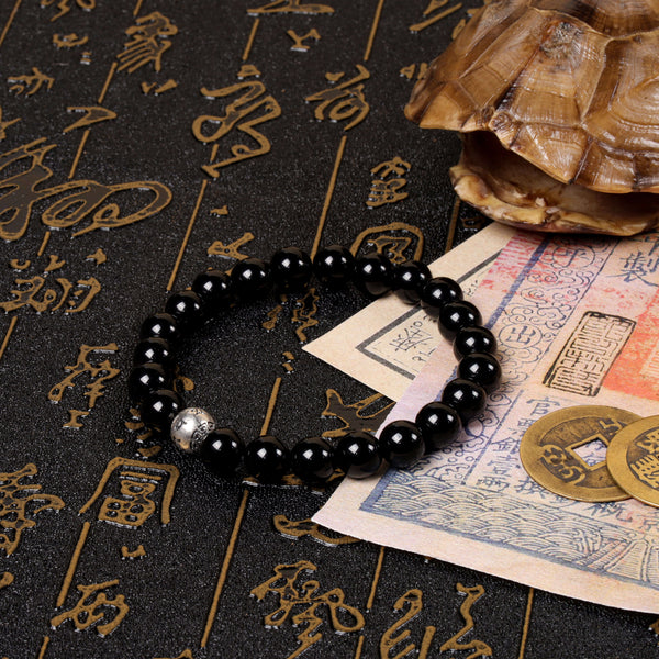 Perspective view of the polished black obsidian bead bracelet with a central silver talisman charm, arranged on a vintage manuscript near a traditional turtle shell and ancient copper cash coins.
