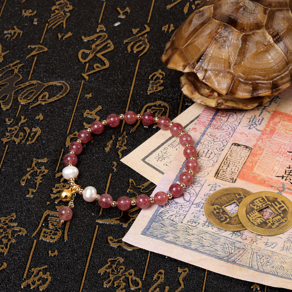 Side perspective view of the delicate pink strawberry quartz talisman bracelet laid over a traditional Chinese manuscript scroll next to ancient copper coins and a textured turtle shell artifact.