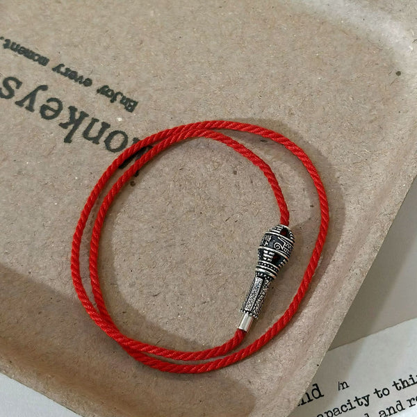 Top-down flat lay view of the red string talisman bracelet on a minimalist card background, highlighting the high-purity sterling silver prayer wheel charm with sacred engravings.